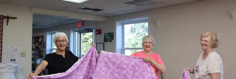 three women hold up a piece of pink fabric