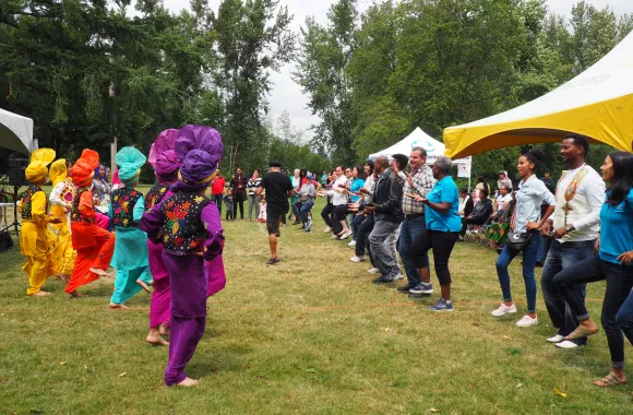 A large group of people gather in a semi circle on grass. Some are dressed in colorful traditional outfits.