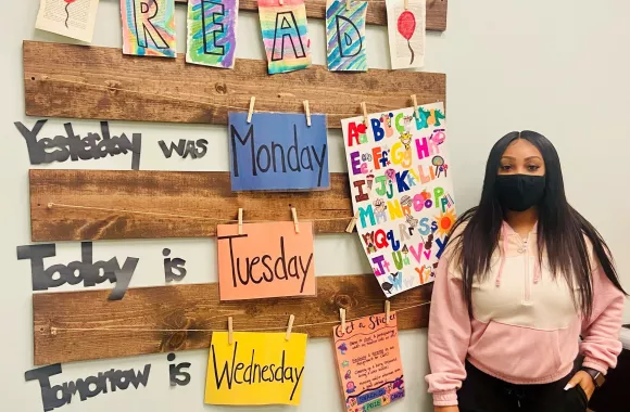 A young woman in a face mask stands next to a wall covered with colorful learning materials in a classroom