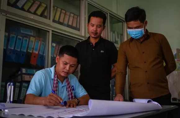 A man sits at a table and writes with a marker onto a large piece of paper while two men stand by and watch