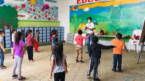 A young man wearing a white polo, a black cap, a face mask and a whistle around his neck stands in front of a group of children leading them in an activity.