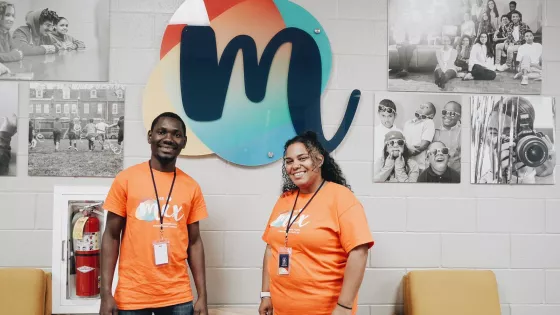 A young man and woman wearing orange t-shirts stand in front of a wall where black and white photos hang as well as a large "M" logo.