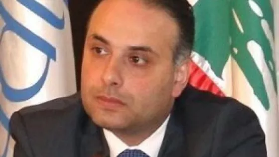 A man wearing a black suit and blue tie sits in front of two flags. 