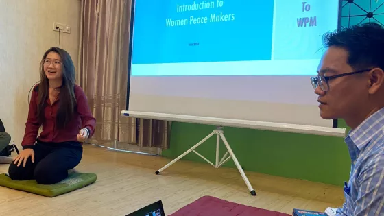 Two people sit on the floor in front of a projector screen. Words on the screen reads, "Introduction to women peace makers."