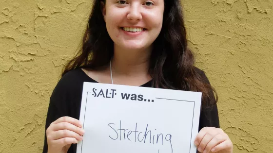 A young adult stands in front of a yellow wall and holds a sign that says, "SALT was stretching"