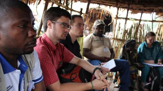 A group of people sit in a hut listening to someone speaking out of view.
