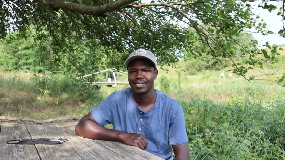 a young man sits at a picnic table outside and smiles at the camera