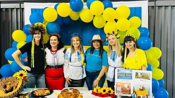 Six women wearing ethnic attire stand in front of an arc of blue and yellow balloons.