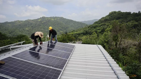 two workers install solar panels on a roof surrounded by mountain views