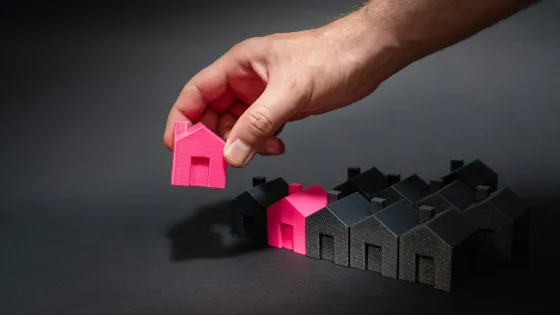 A hand holds up a pink model of a house from a row of black home models.