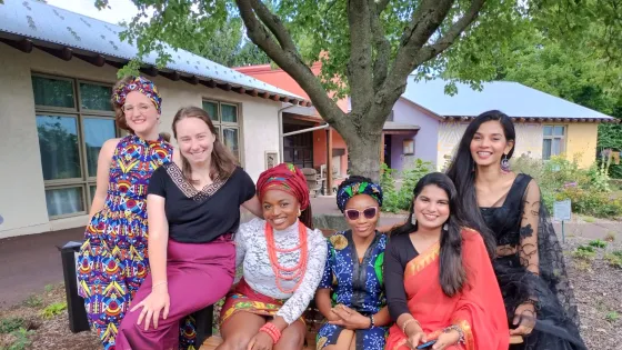 A group of five people wearing traditional clothing representing different cultures sit together in front of a tree.