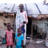 Mary Nyashin Tsief (70) stands in front of her home in Rubkona camp for people displaced by flooding and violent conflict. She is accompanied by her two grandchildren, Nyamen Iuok Ram (age 5) and Nyawich Iuok Ram (age 3), who live in a tent adjacent to hers.